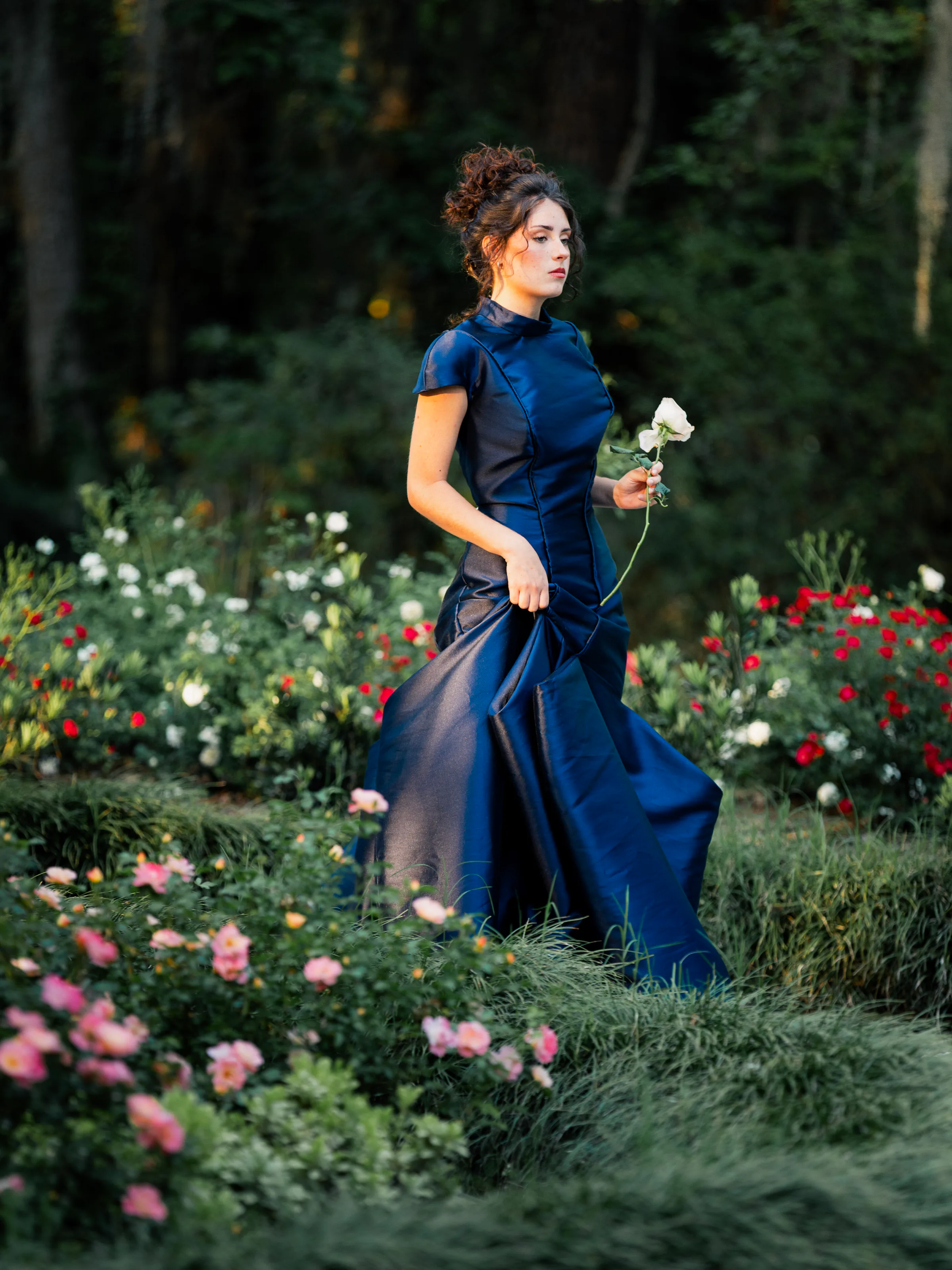 Vesper navy satin gown moving through a garden with gathered skirt panels and curved bodice seams