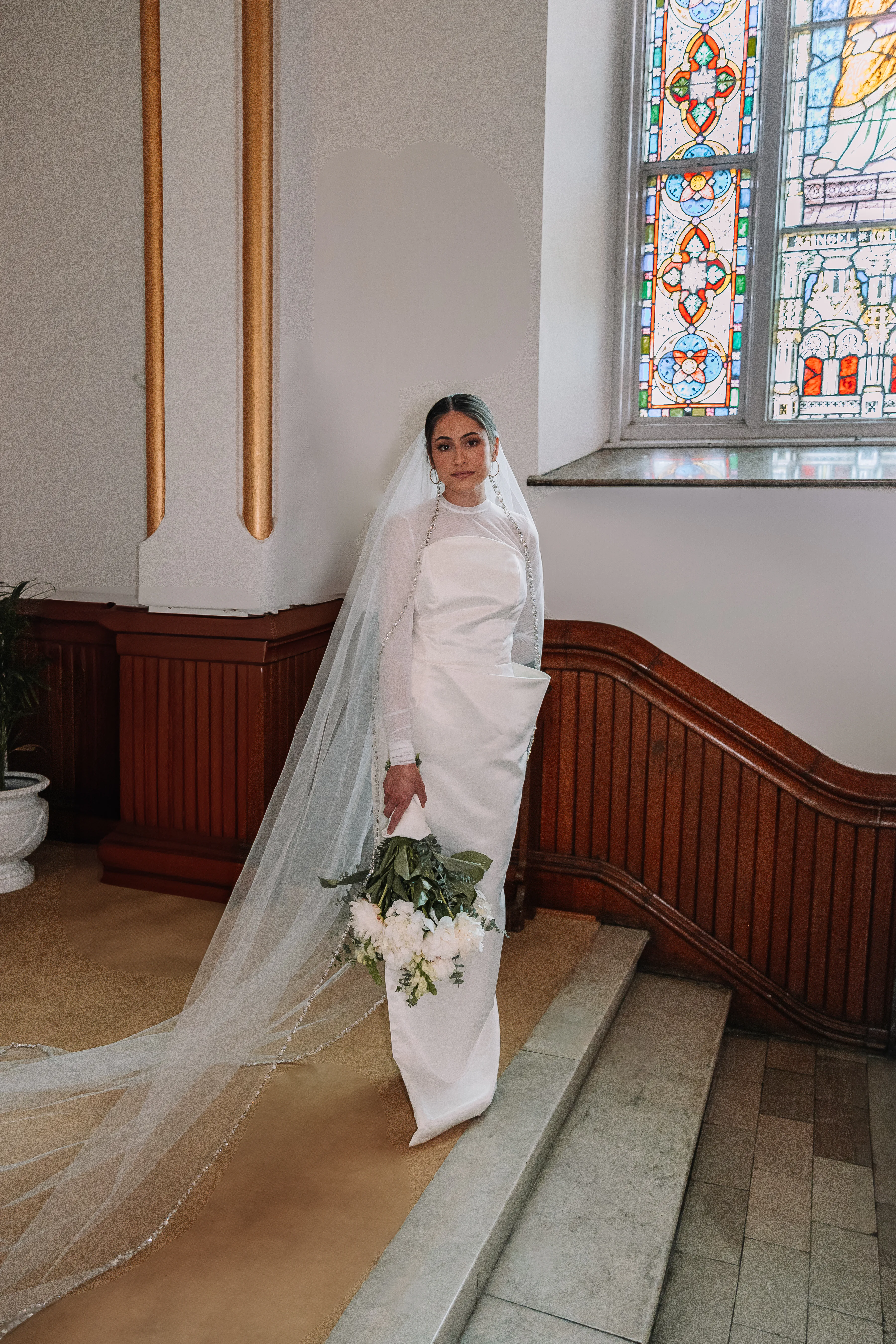 A bride wearing Samantha Covey's asymmetrical wedding dress with a long veil beside stained glass windows.