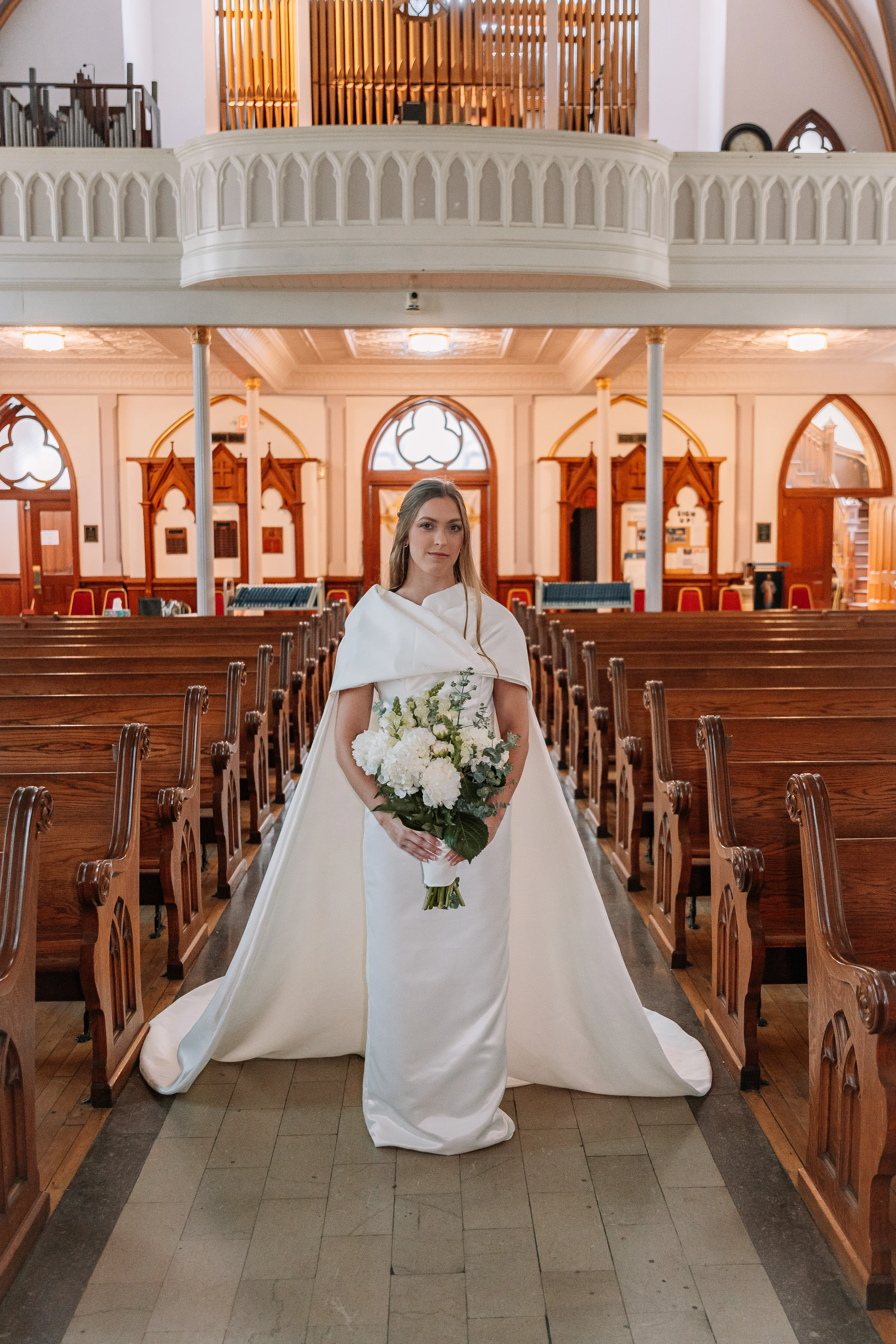 A bride wearing Samantha Covey's detachable cape wedding dress standing in a church aisle with a white bouquet.