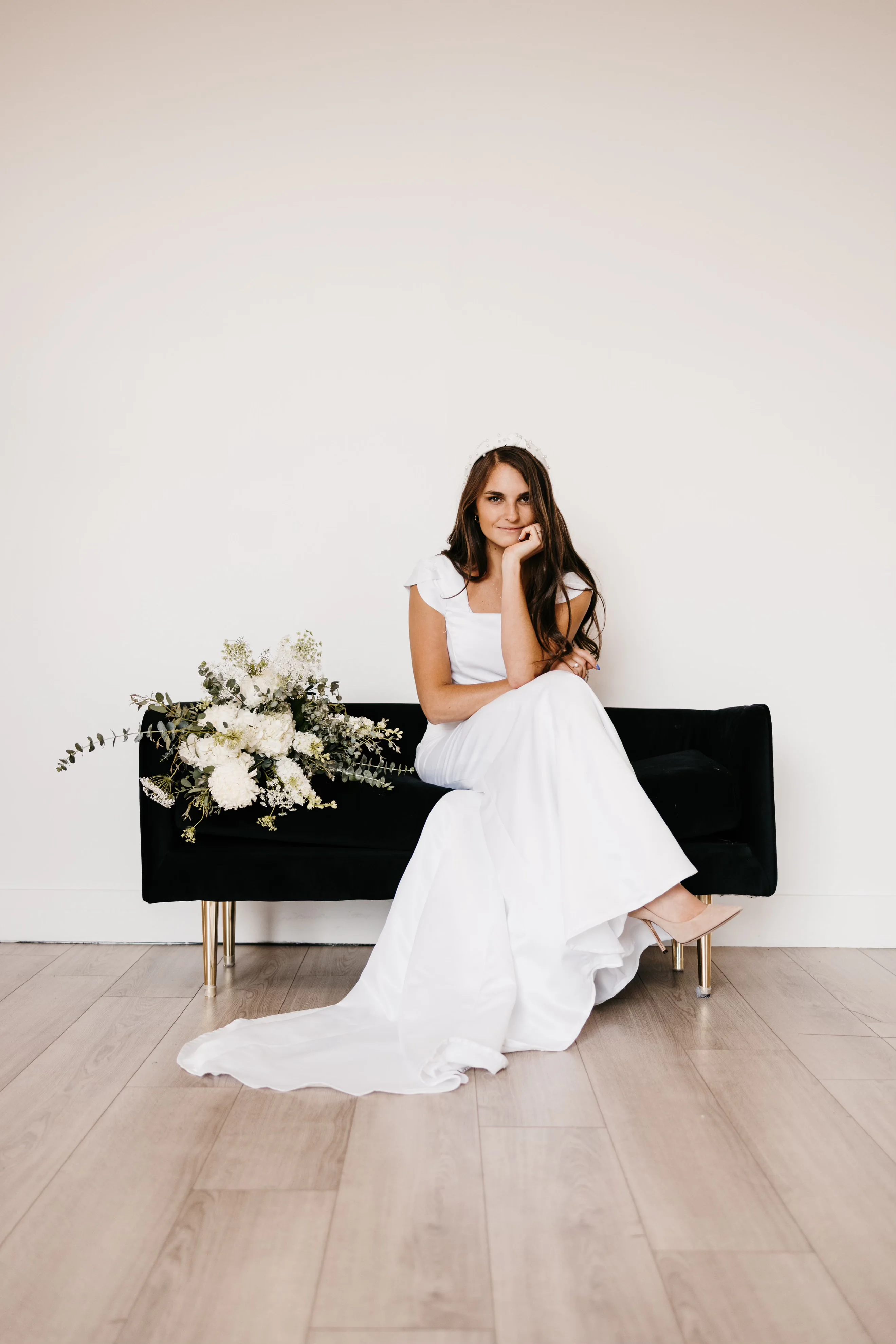 A seated front portrait of the first wedding dress on a black settee beside a white bouquet.
