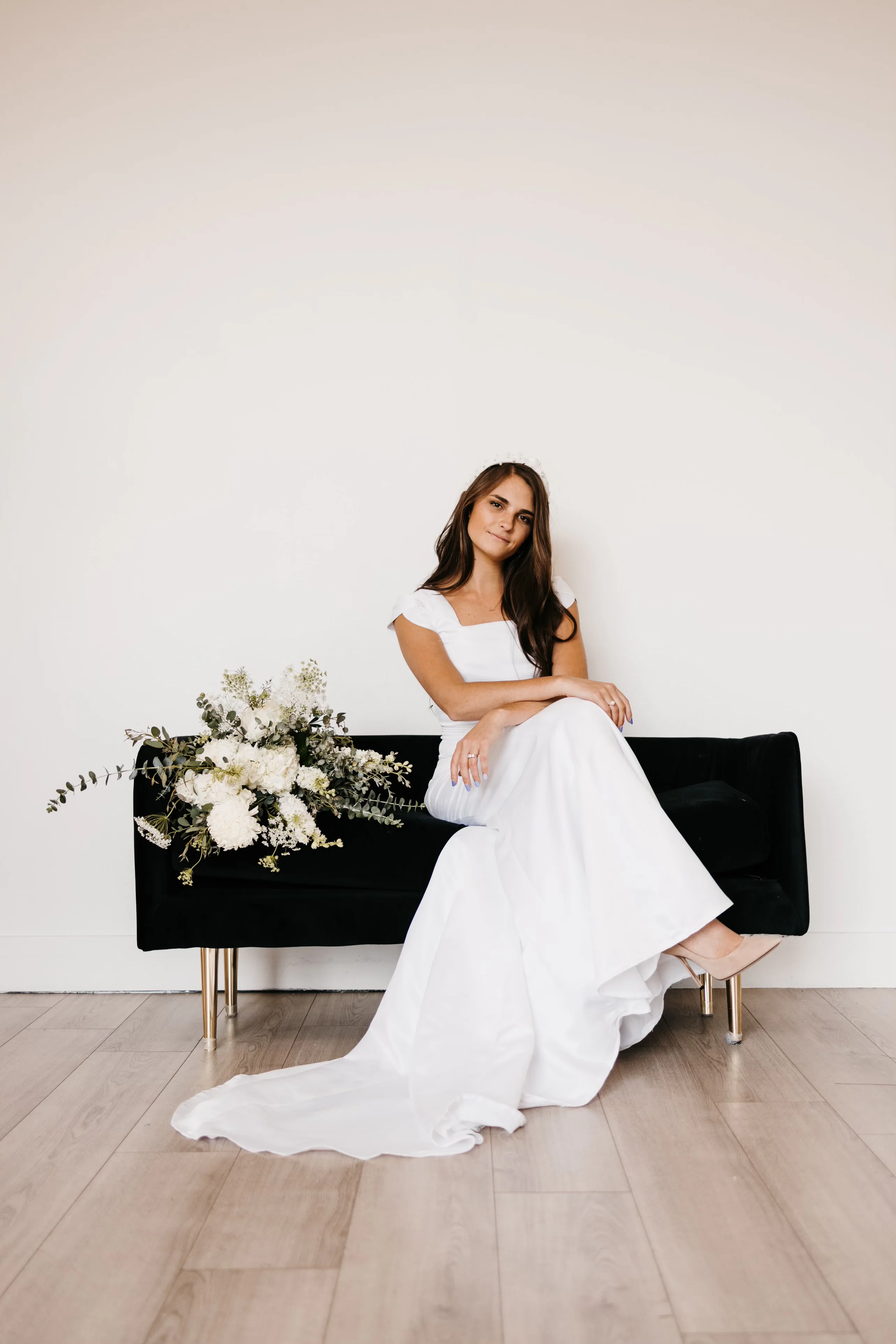 A seated bridal portrait on a black settee, showing the long skirt and soft square neckline of the first wedding dress Samantha Covey made.