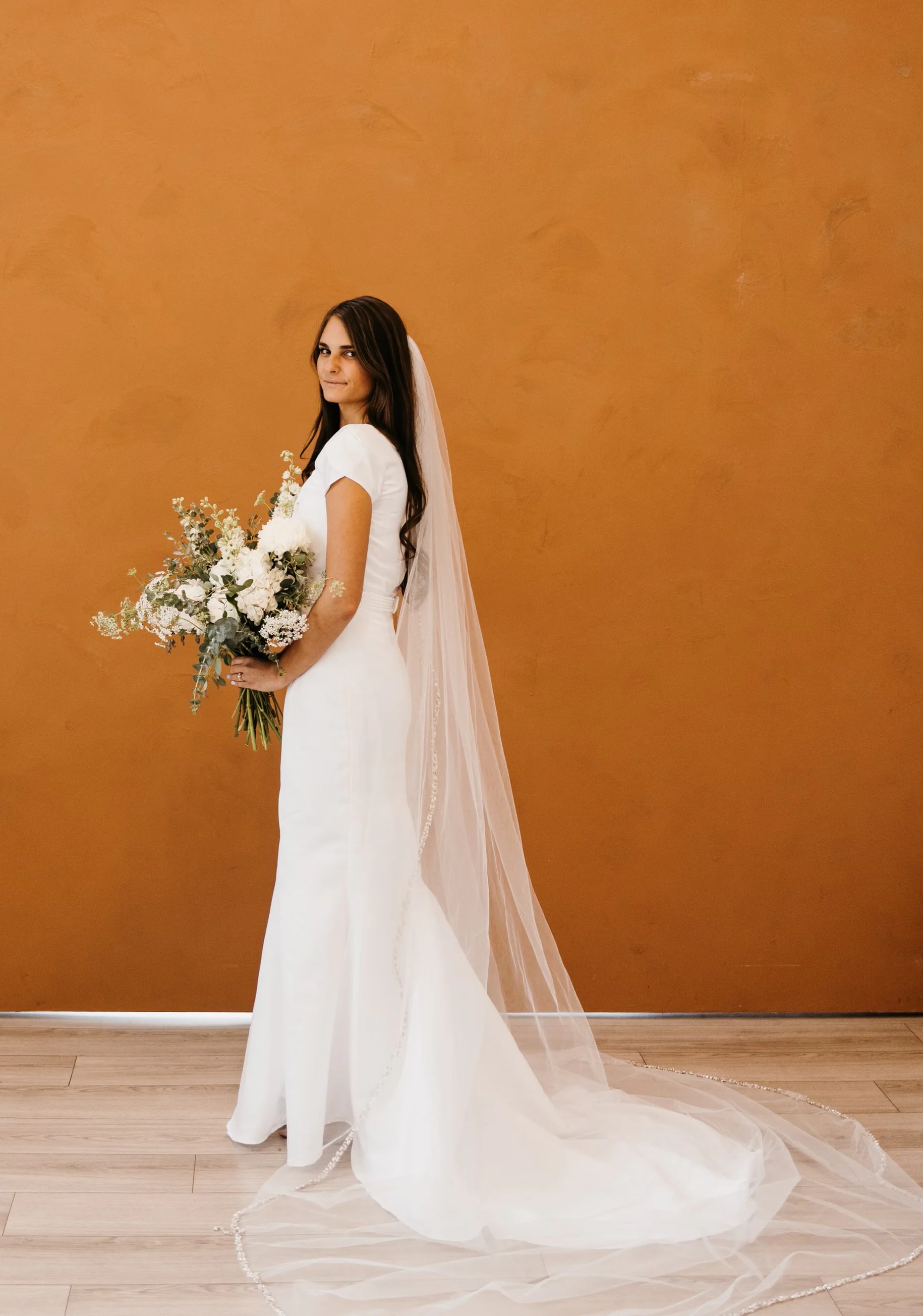 A side portrait of the first wedding dress with a white bouquet and long veil against a rust-colored wall.