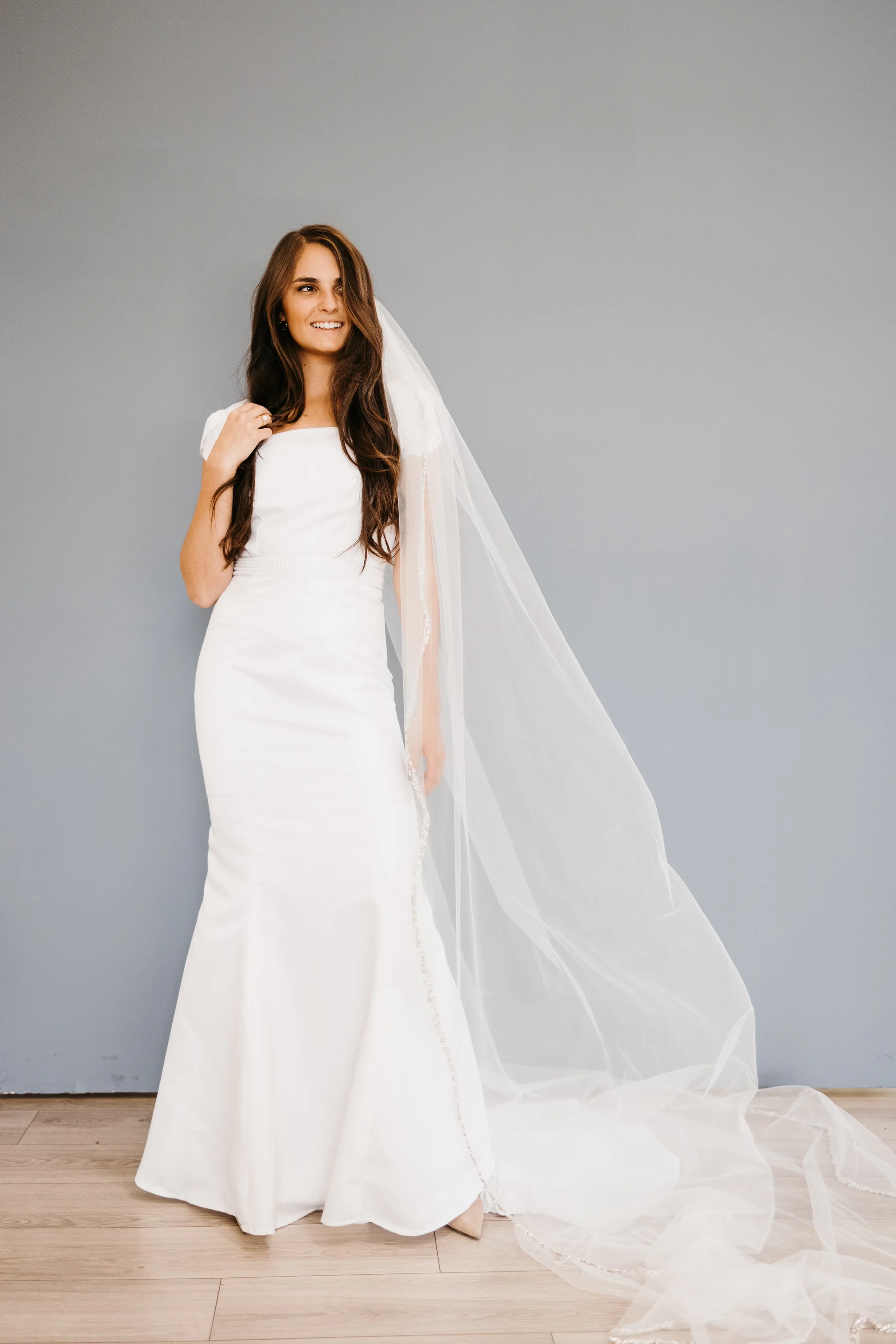 A full-length portrait of the first wedding dress Samantha Covey made, styled with a long veil against a gray wall.