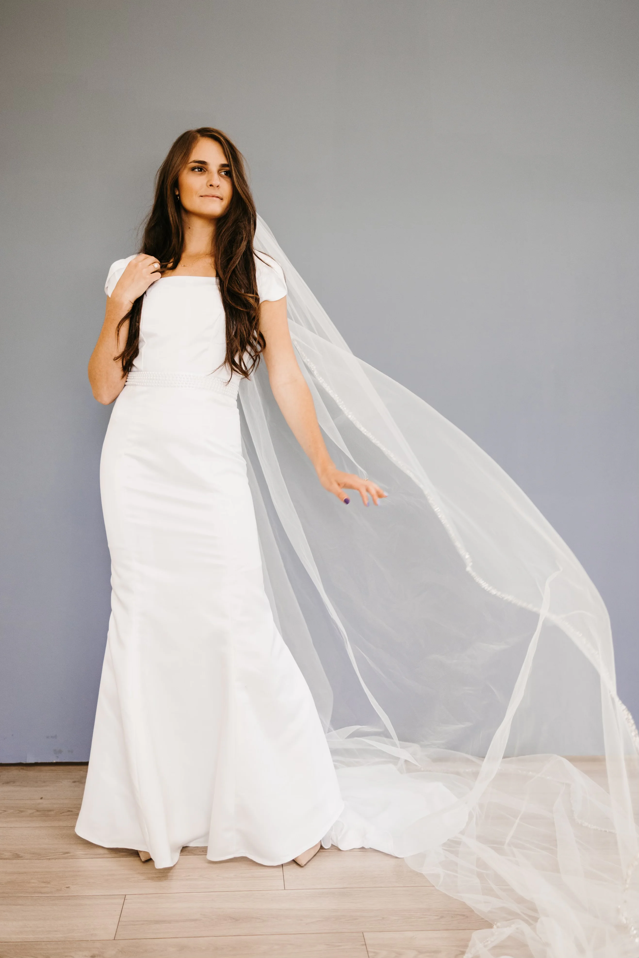 A standing bridal portrait showing the dress with softly shaped sleeves, a fitted waist, and a long veil moving to the side.