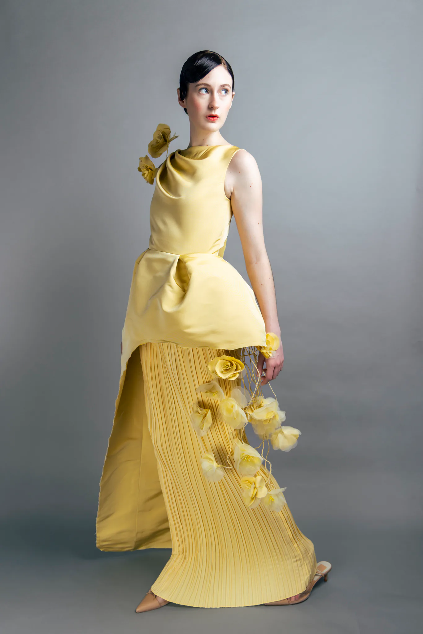A full studio portrait of a model wearing Samantha Covey's yellow sculptural dress with a pleated skirt and cascading handmade flowers.