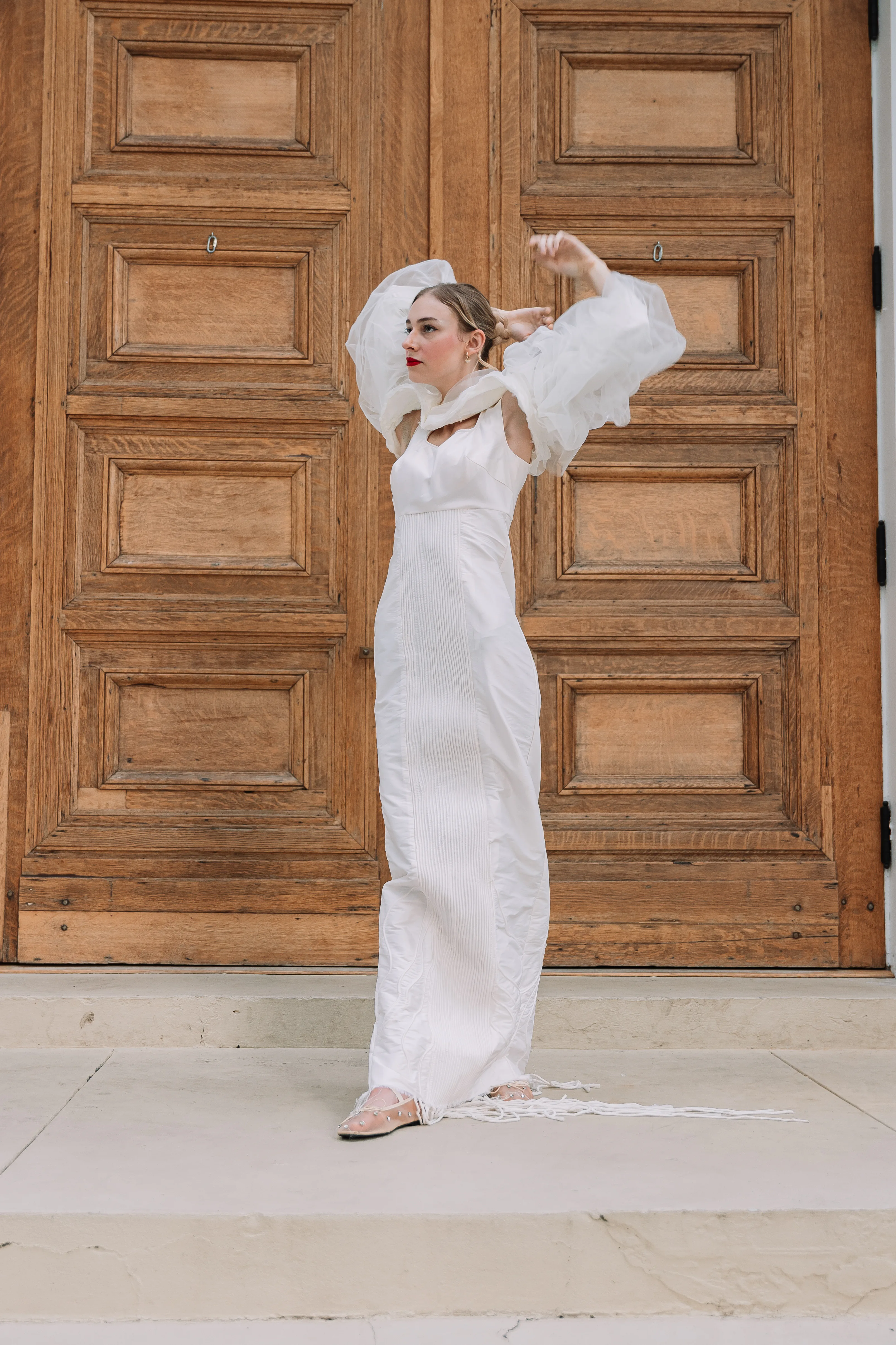 A full-length portrait of The White Project dress in front of carved wood doors, showing the white column silhouette, translucent sleeve volume, and hand-piped front panel.