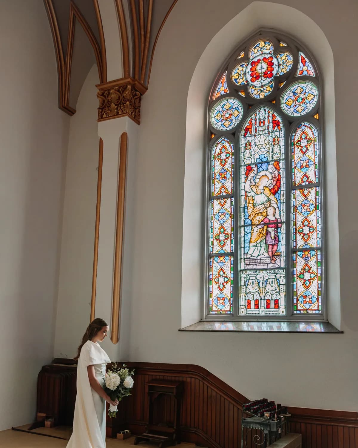 A bride holding a bouquet beneath a tall stained-glass church window.