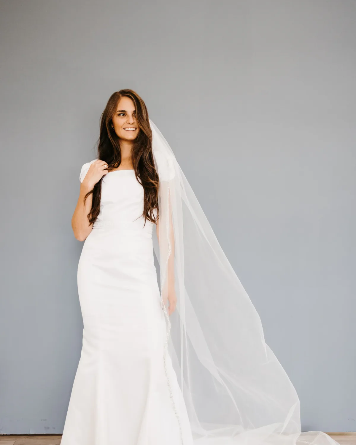 A bride in a simple white gown and veil against a clean gray backdrop.