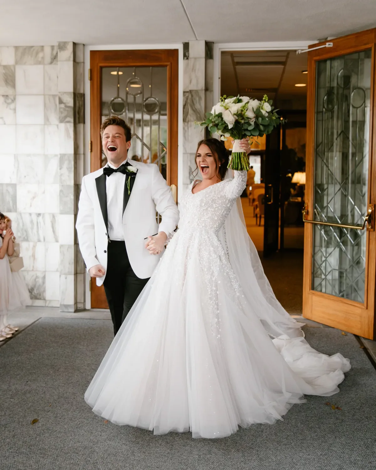 A bride in a white wedding dress with sleeves leaving a church entrance with her groom.