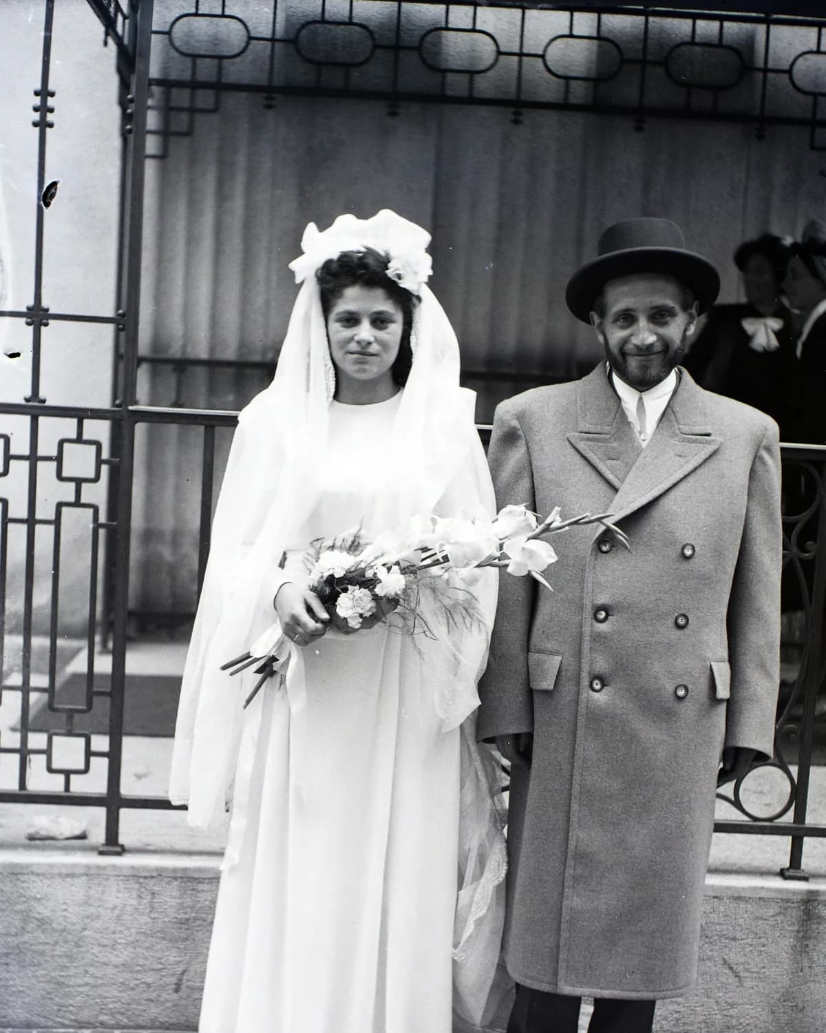 A black-and-white portrait of an Orthodox Jewish bride and groom outside a synagogue.