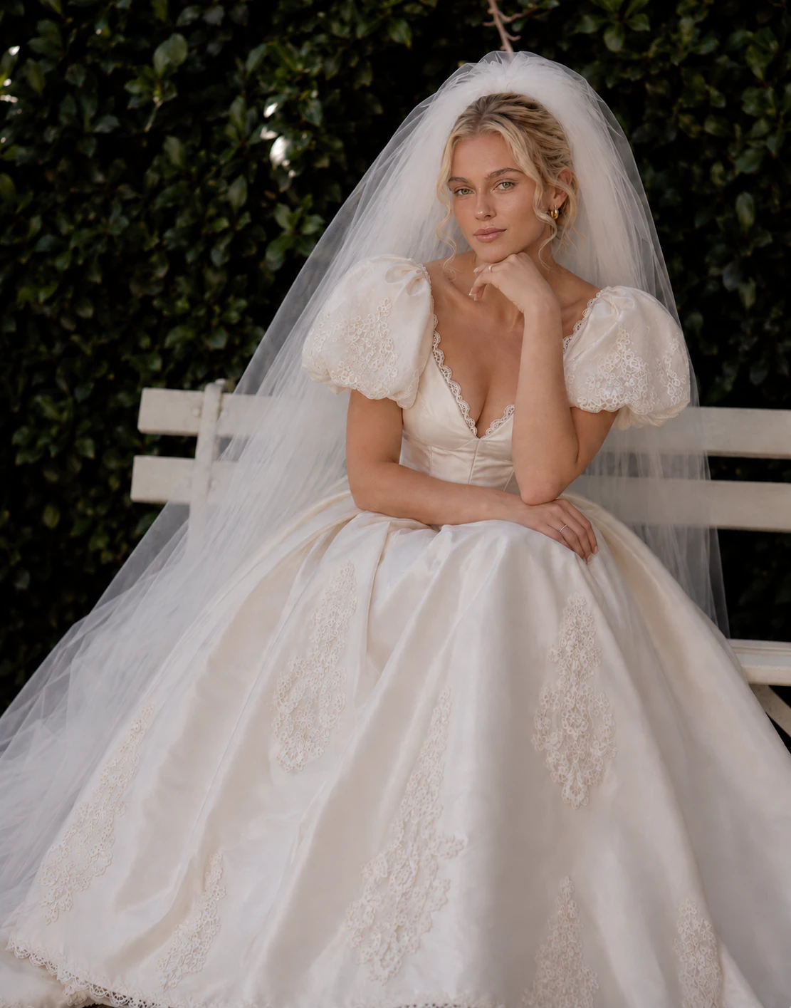 Bride seated in Noelle with a flowing veil, lace-trimmed puff sleeves, and a full satin skirt with lace motifs