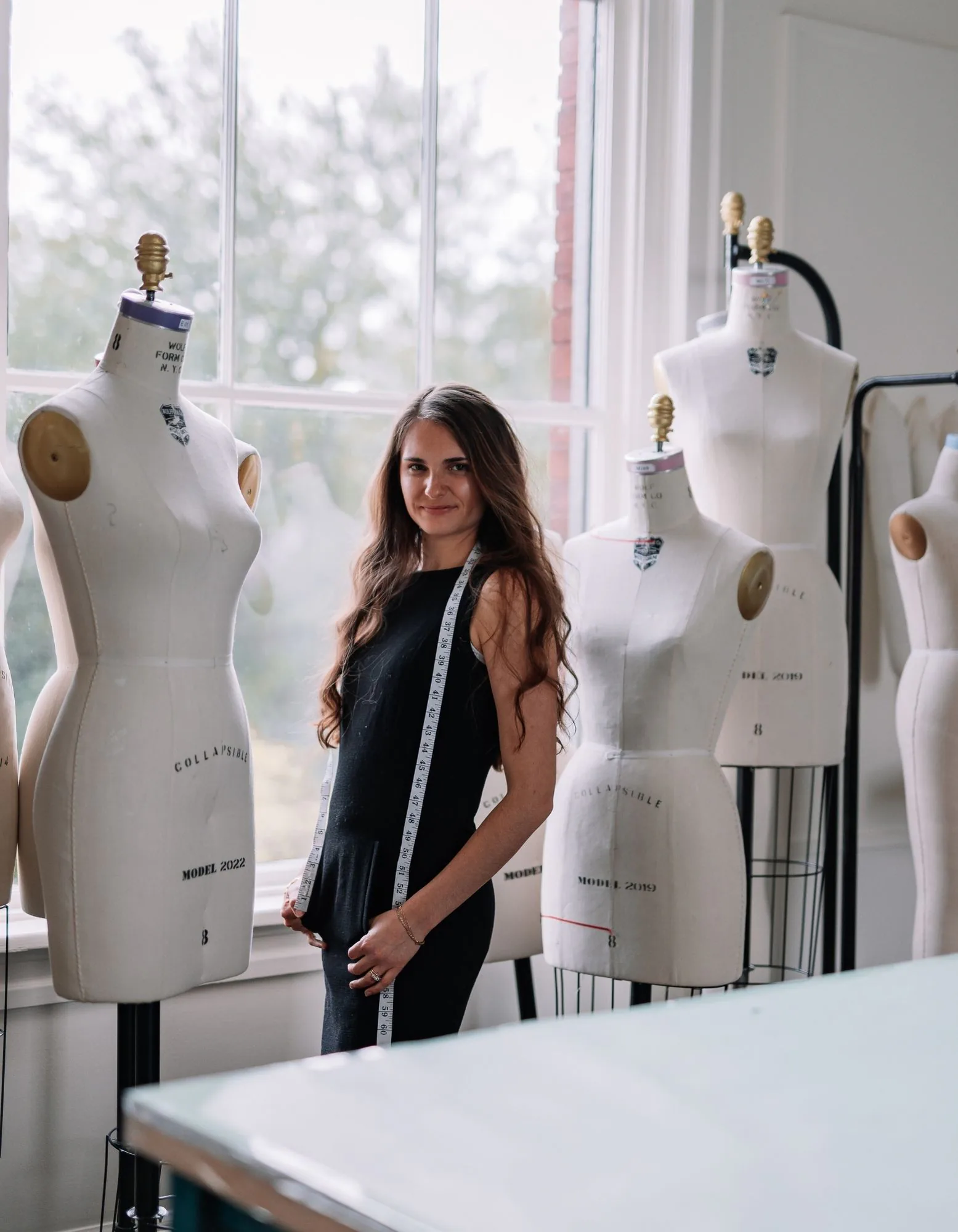 Samantha Covey standing in her atelier among dress forms with a measuring tape around her neck.
