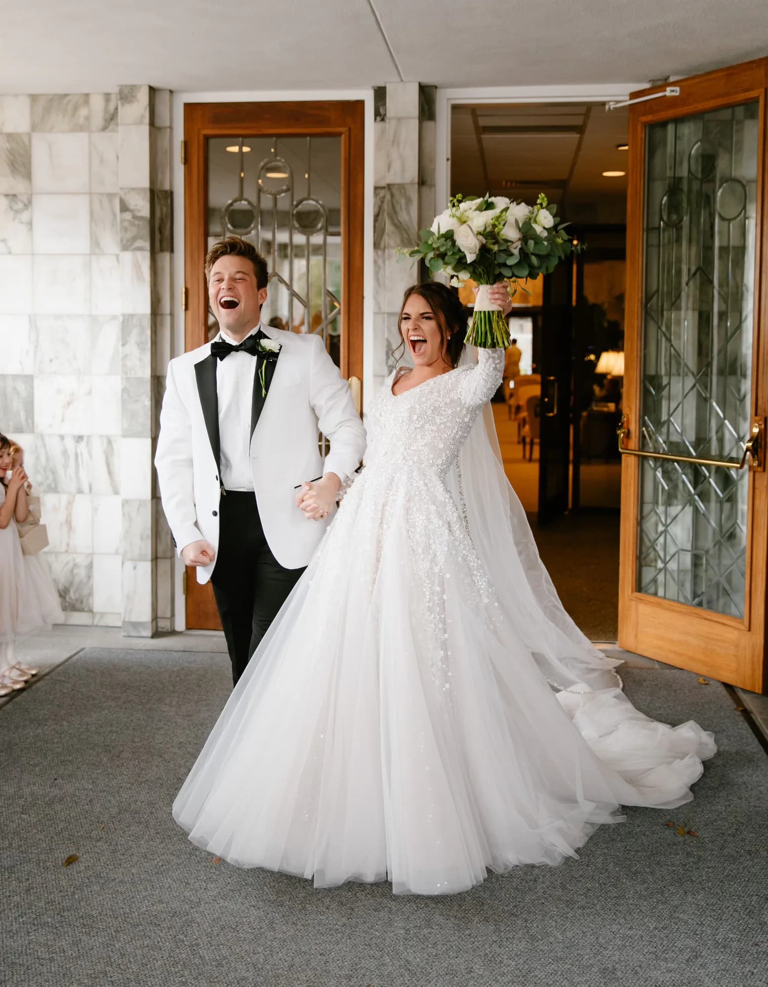 Samantha Covey wedding dress A bride wearing a Samantha Covey wedding dress exits a church holding a white bouquet beside the groom.