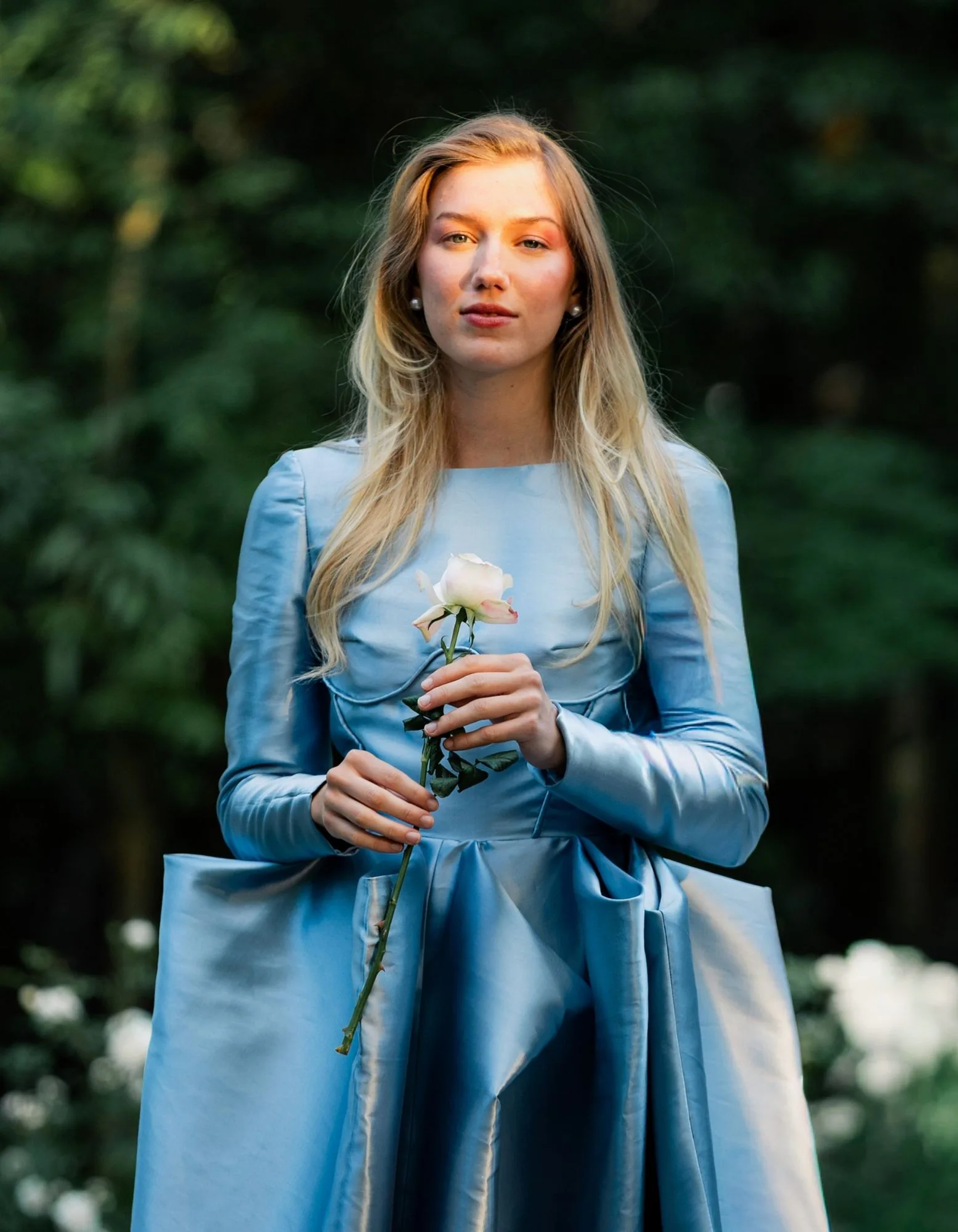 A model in a powder-blue long-sleeve gown holding a pale rose in a wooded garden.