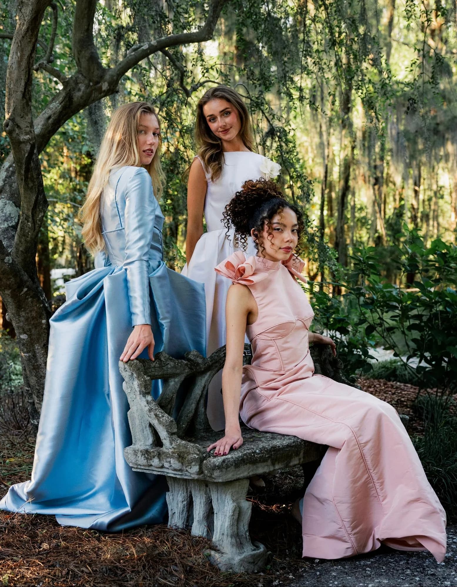 Three models in ivory, powder blue, and blush gowns posed around a stone bench in the garden.