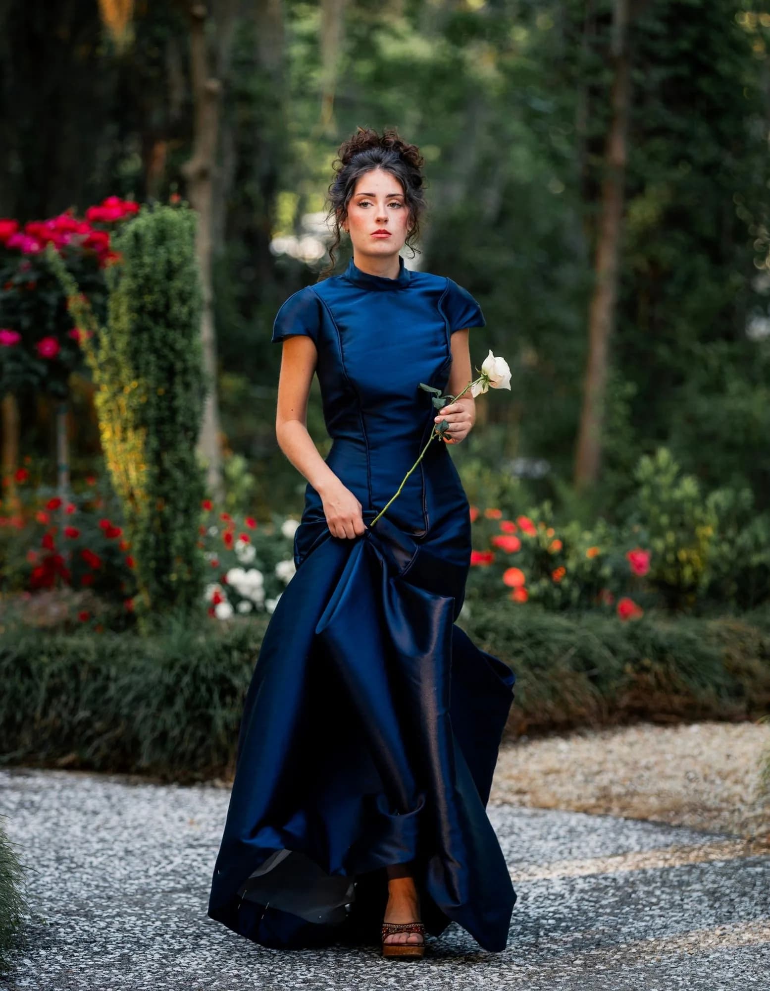 A midnight-blue gown moving through a rose garden with a single white rose.