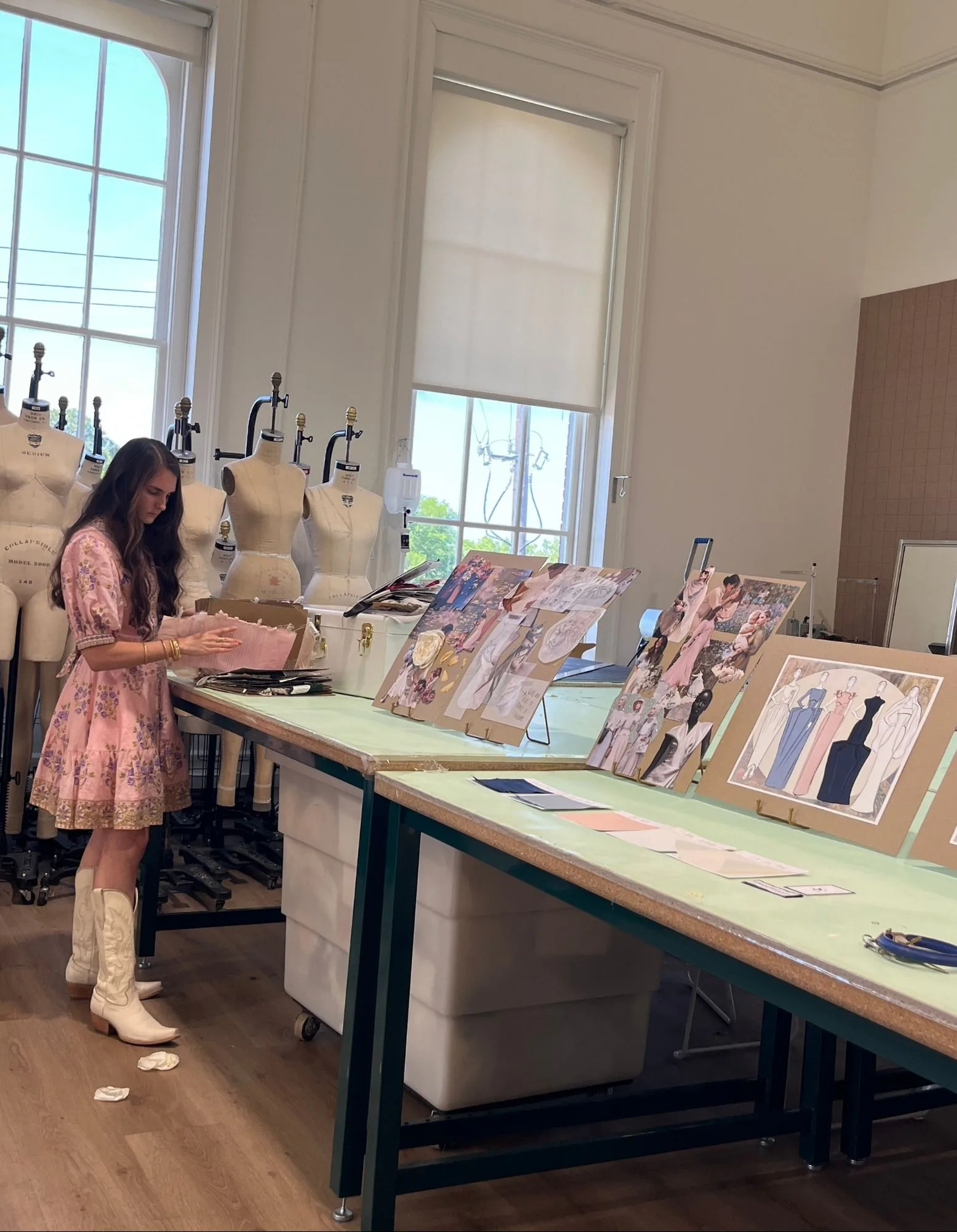 Samantha working at the atelier table with dress forms, sketches, fabric swatches, and moodboards.