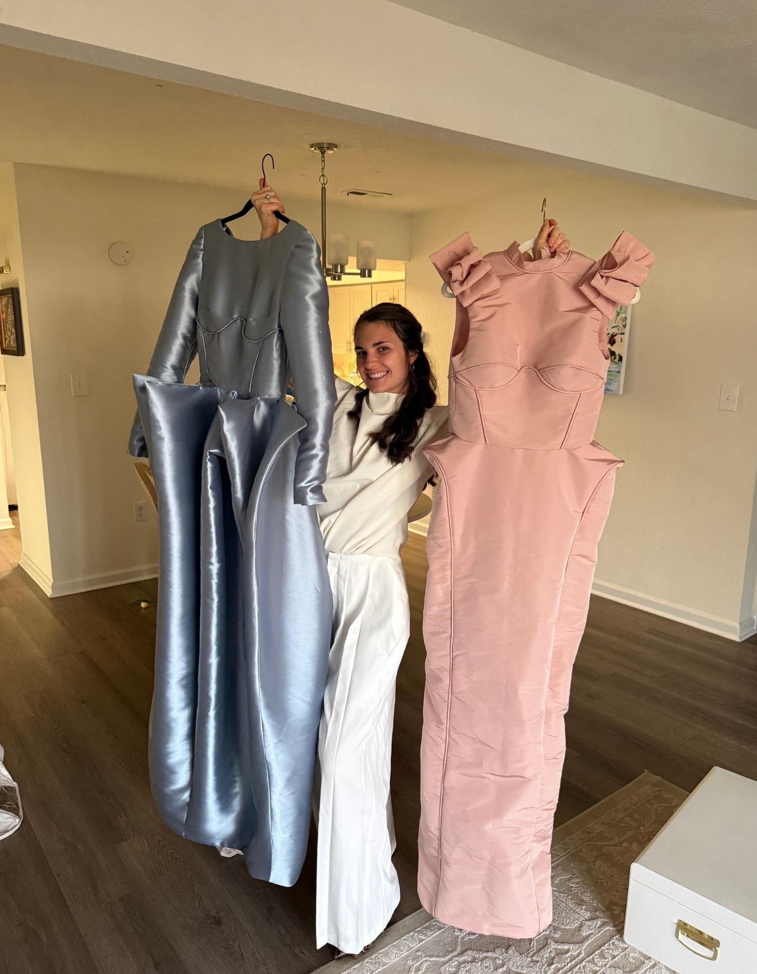 Samantha smiling while holding the powder-blue satin gown and blush bow-shoulder gown on hangers.