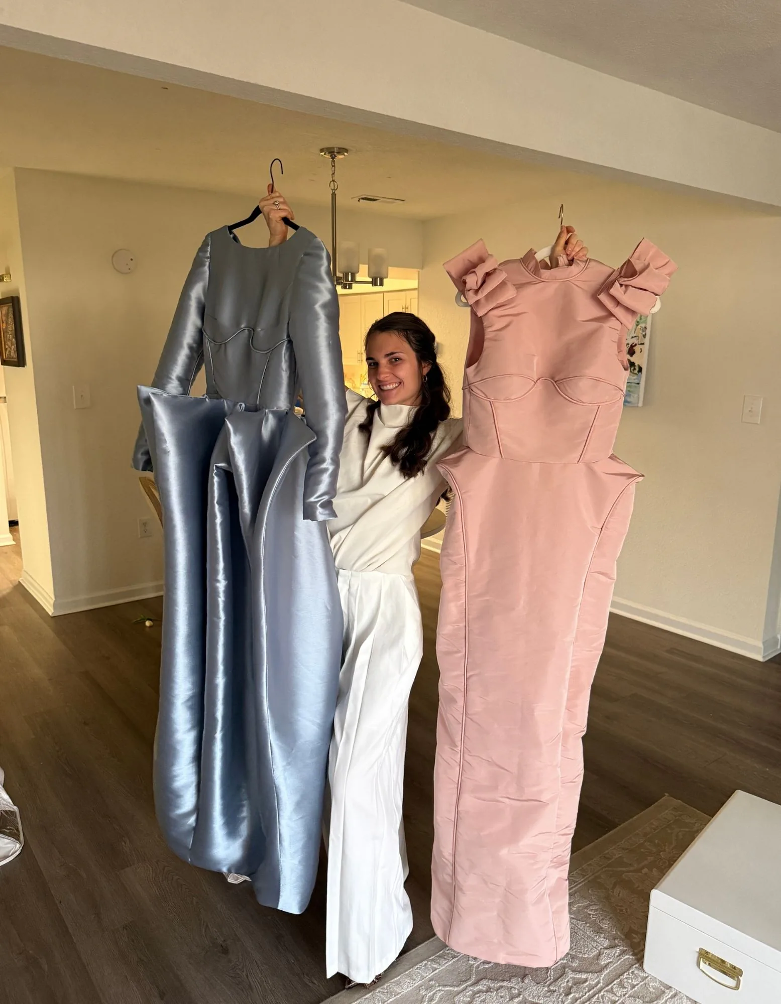 Samantha smiling while holding the powder-blue satin gown and blush bow-shoulder gown on hangers.