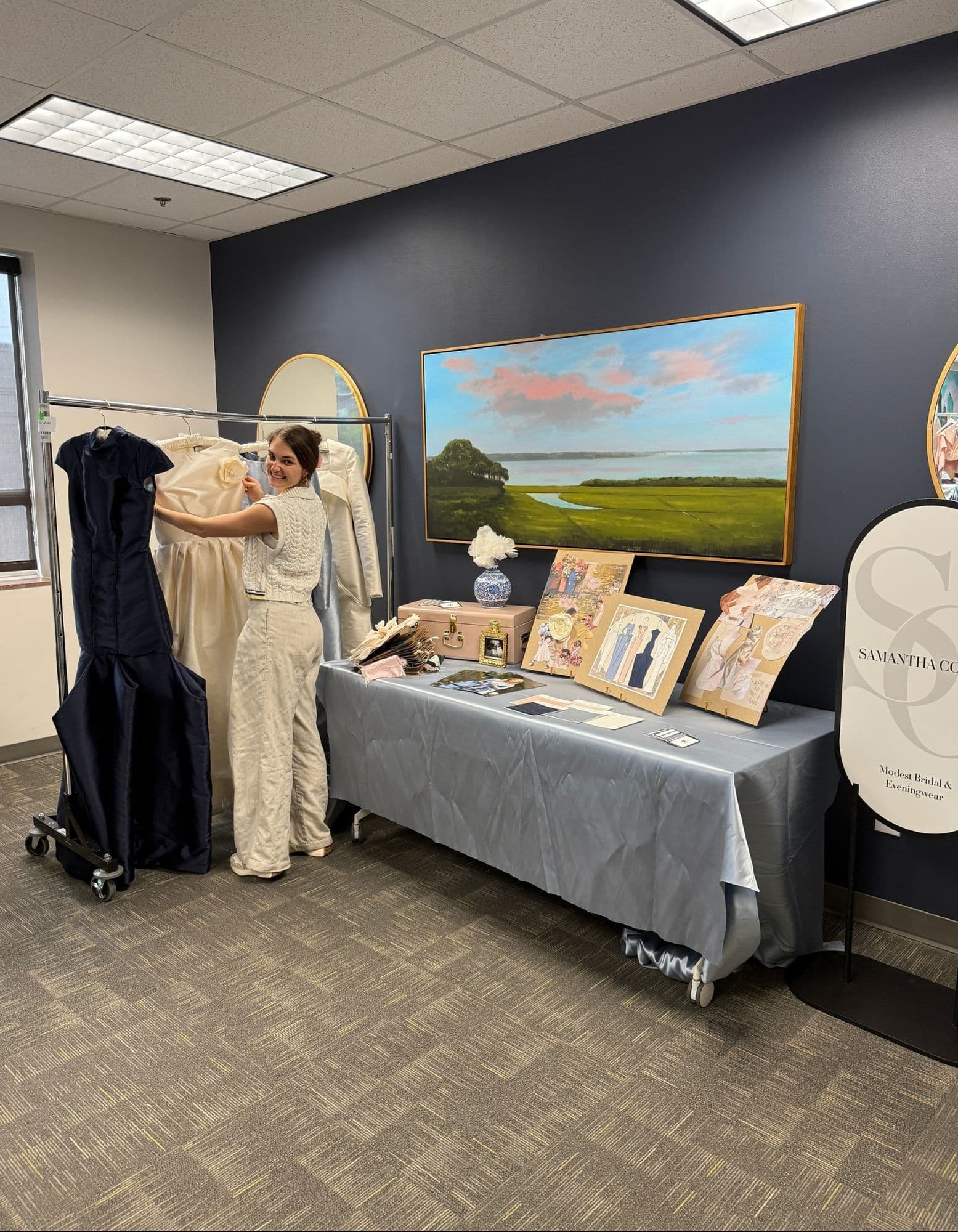 Samantha arranging ivory and navy gowns beside a display of sketches, swatches, moodboards, and Samantha Covey signage.
