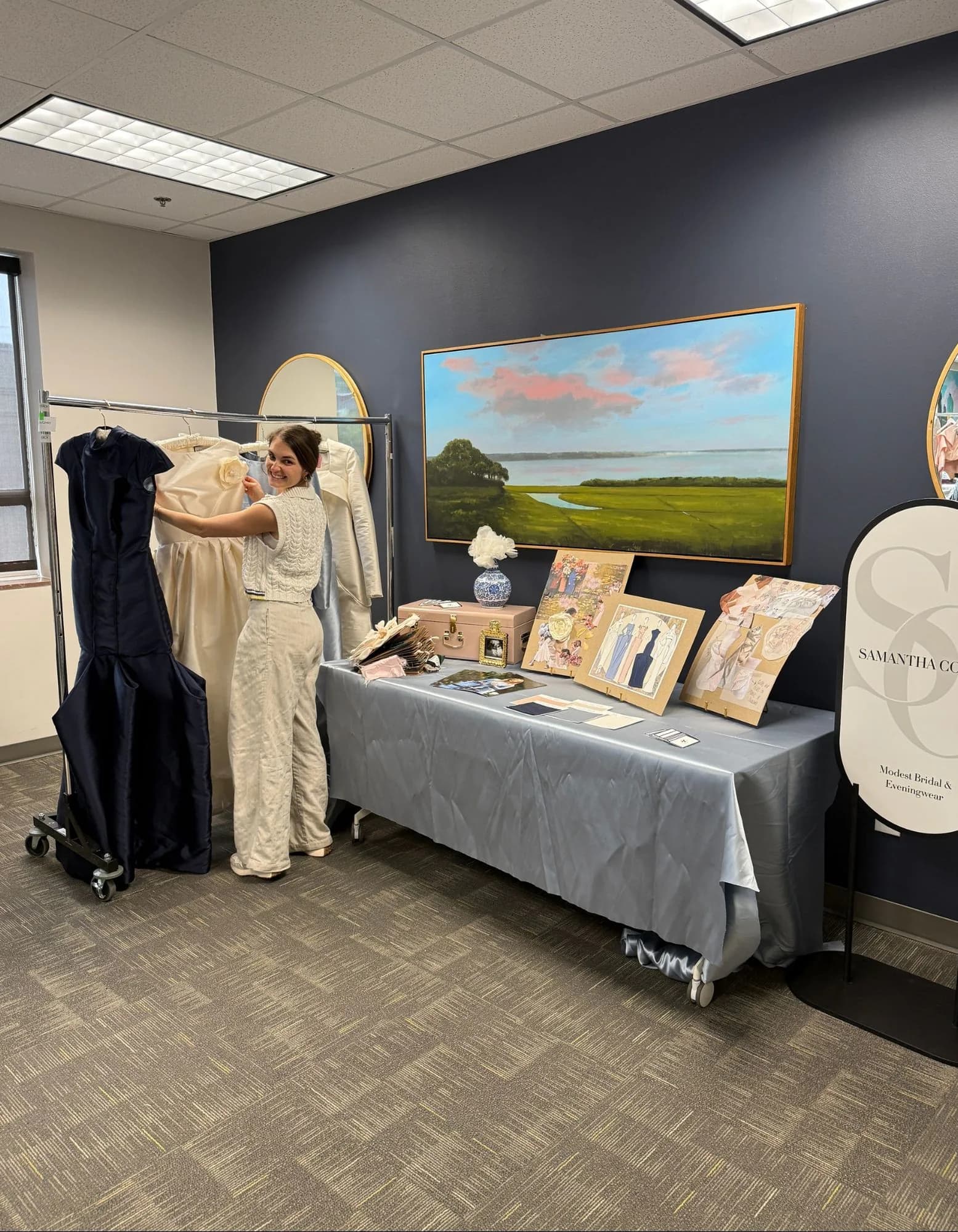 Samantha arranging ivory and navy gowns beside a display of sketches, swatches, moodboards, and Samantha Covey signage.