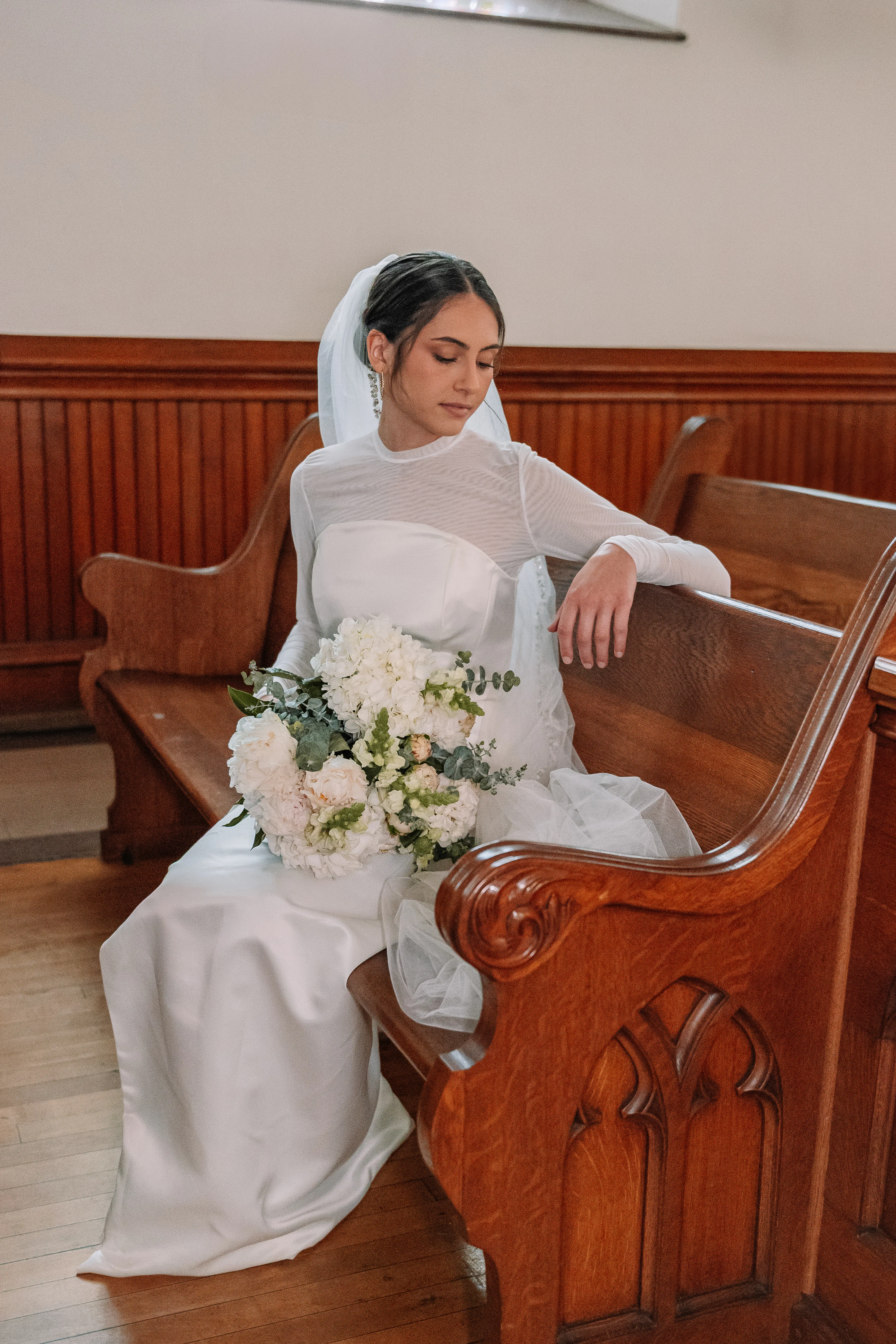 A bride in the asymmetrical wedding dress seated in a wooden church pew with a white bouquet.