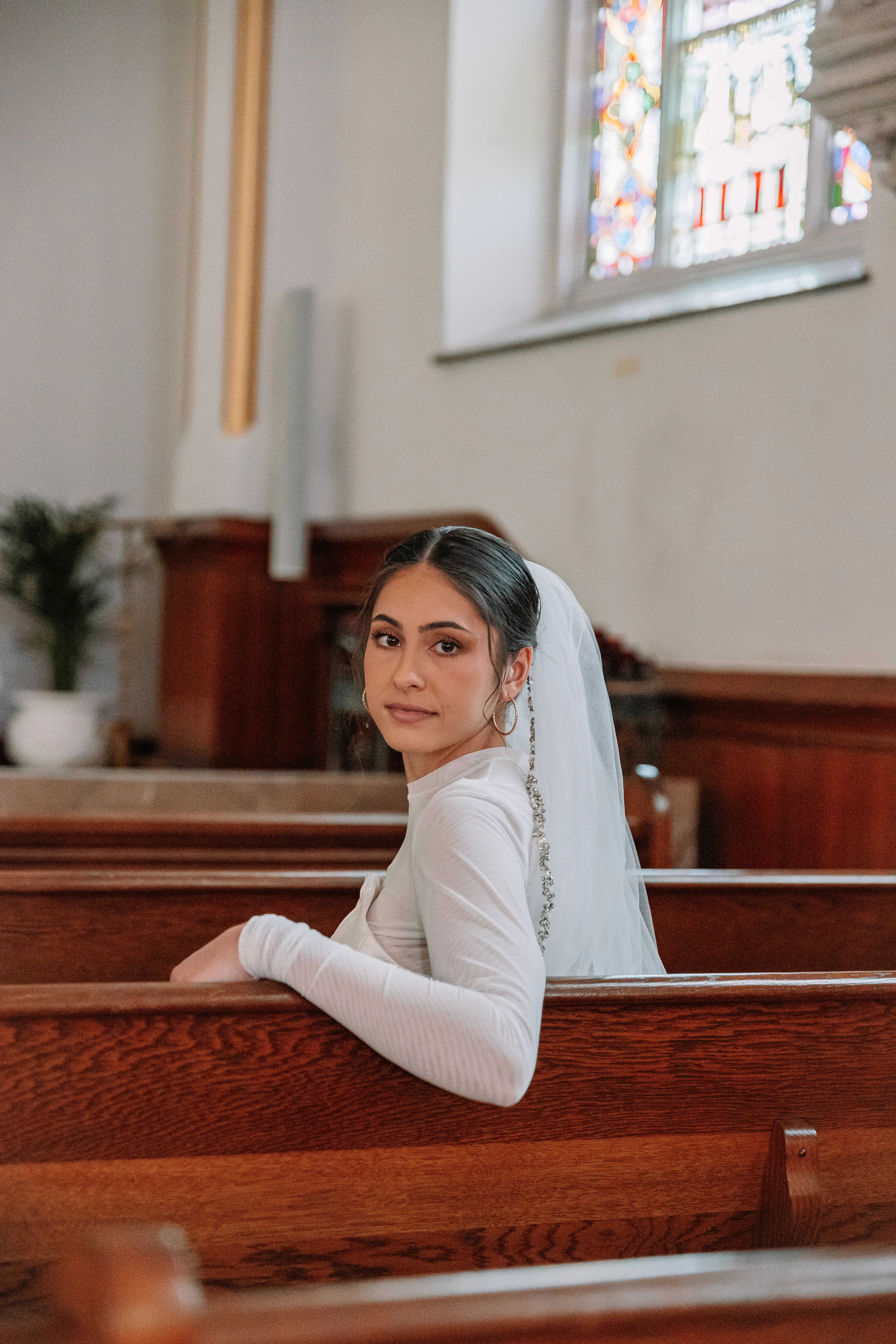 A bride in the asymmetrical wedding dress turning back from a wooden church pew.