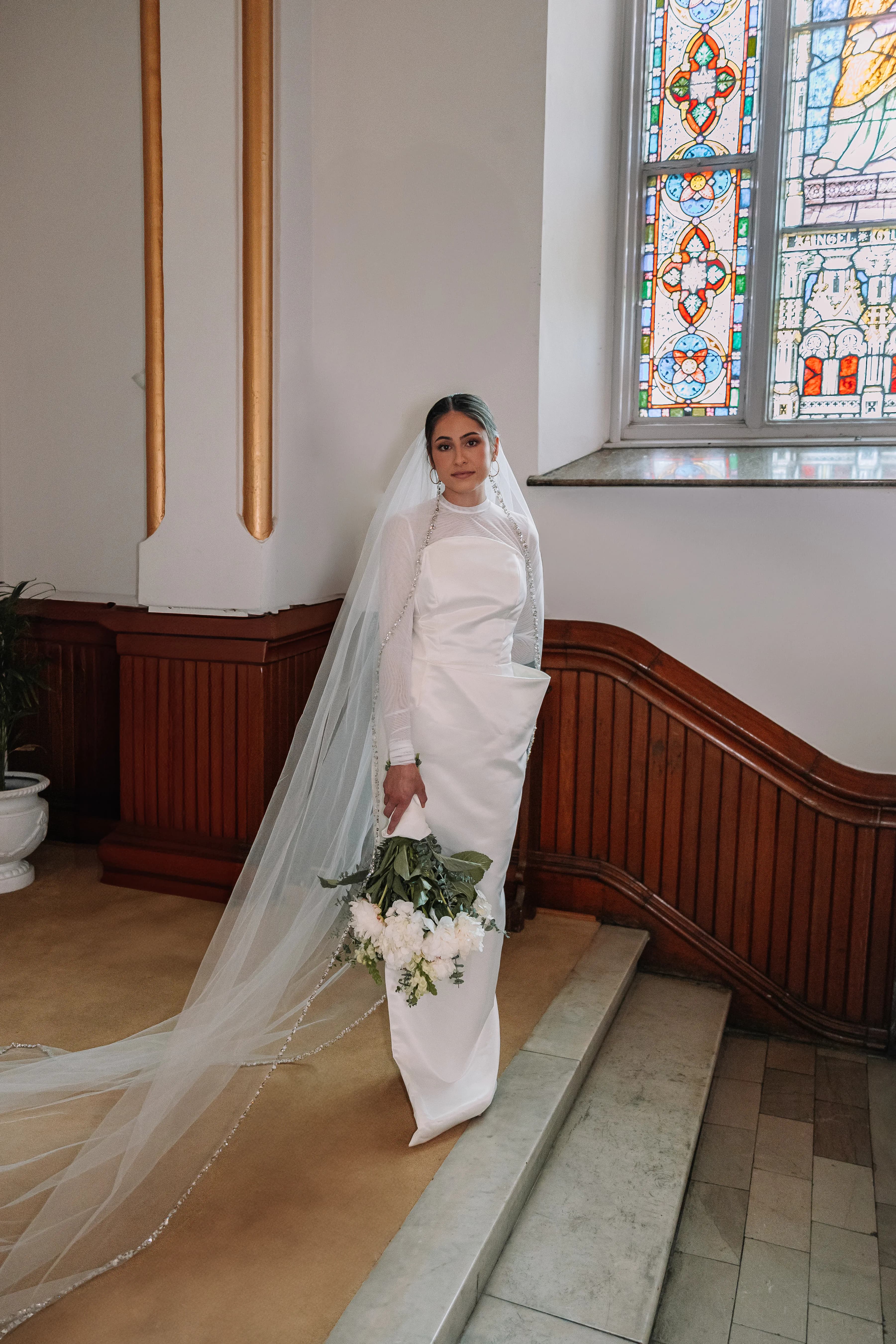 A bride wearing Samantha Covey's asymmetrical wedding dress with a long veil beside stained glass windows.