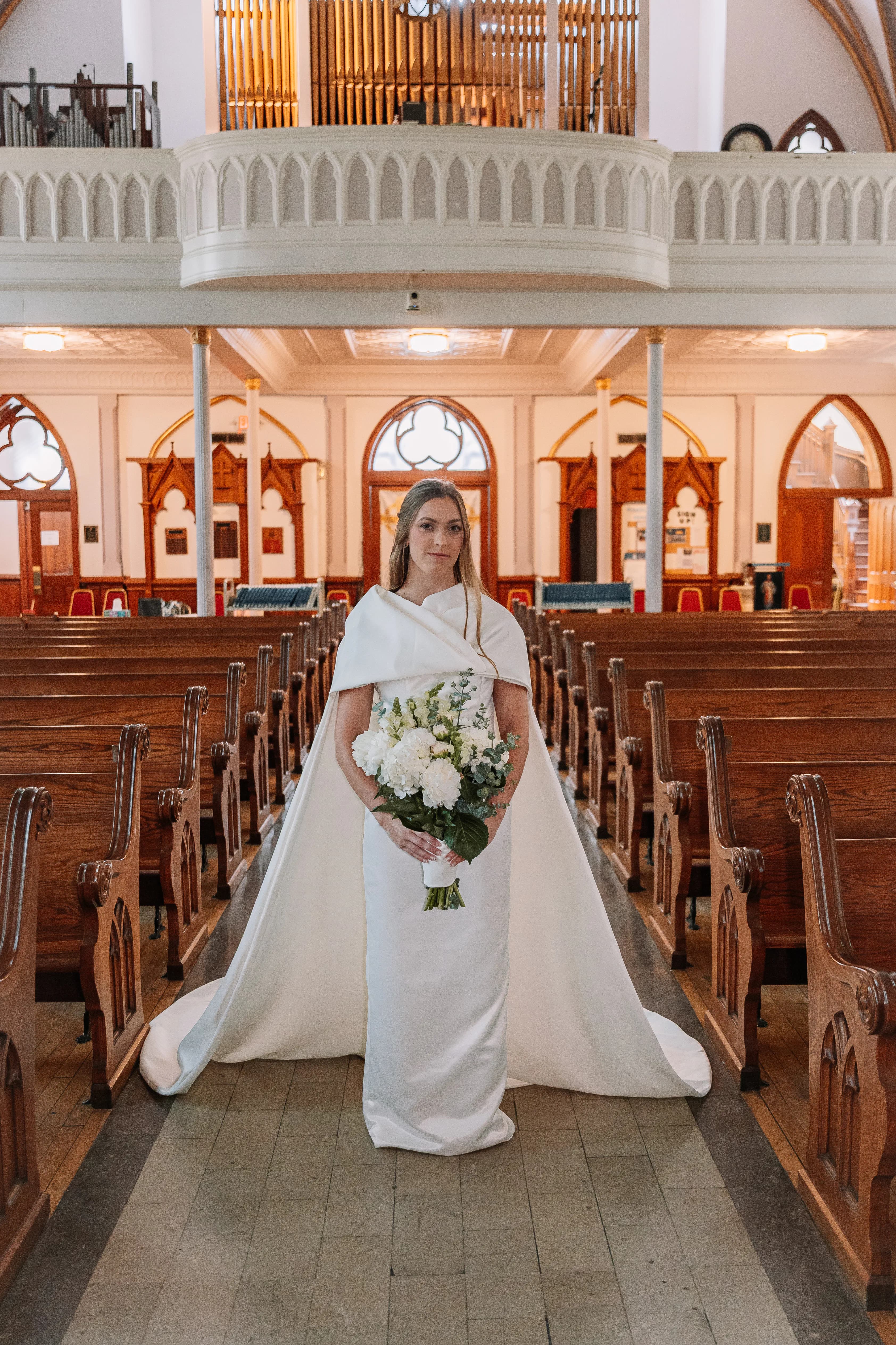 A bride wearing Samantha Covey's detachable cape wedding dress standing in a church aisle with a white bouquet.