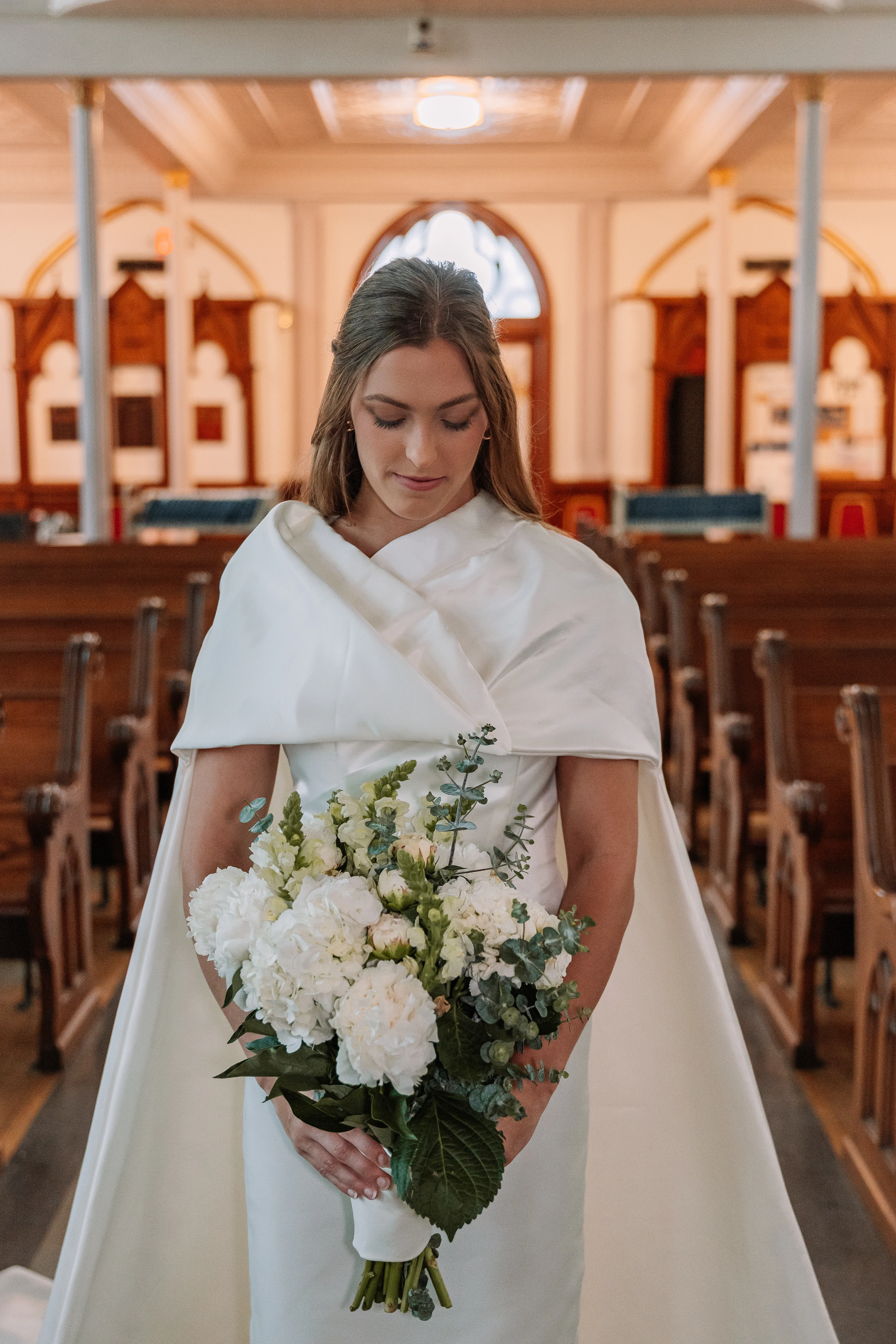 A closer portrait of the detachable cape wedding dress with the bride holding a white bouquet in the aisle.