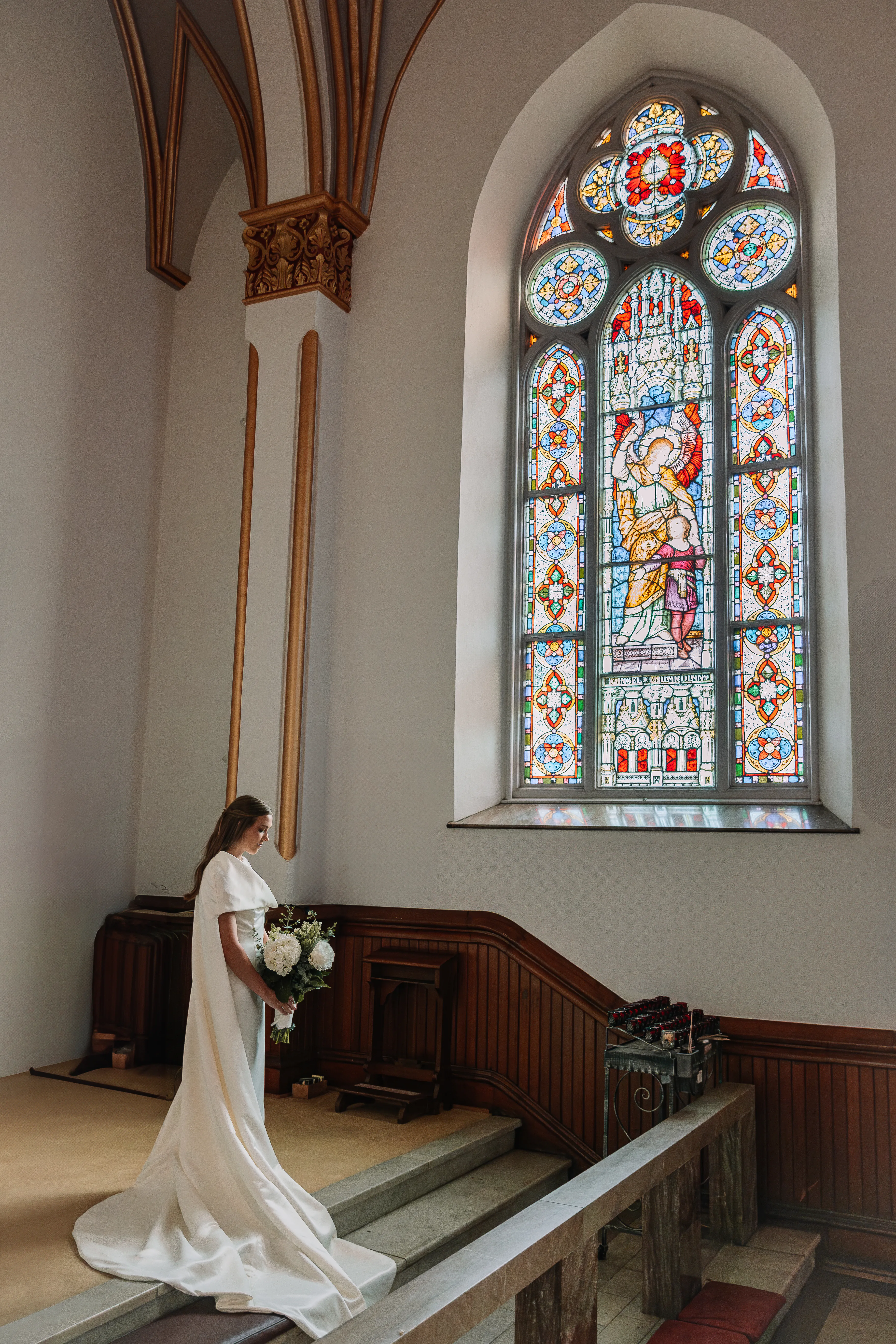 A full cathedral portrait of the detachable cape wedding dress beneath a tall stained glass window.