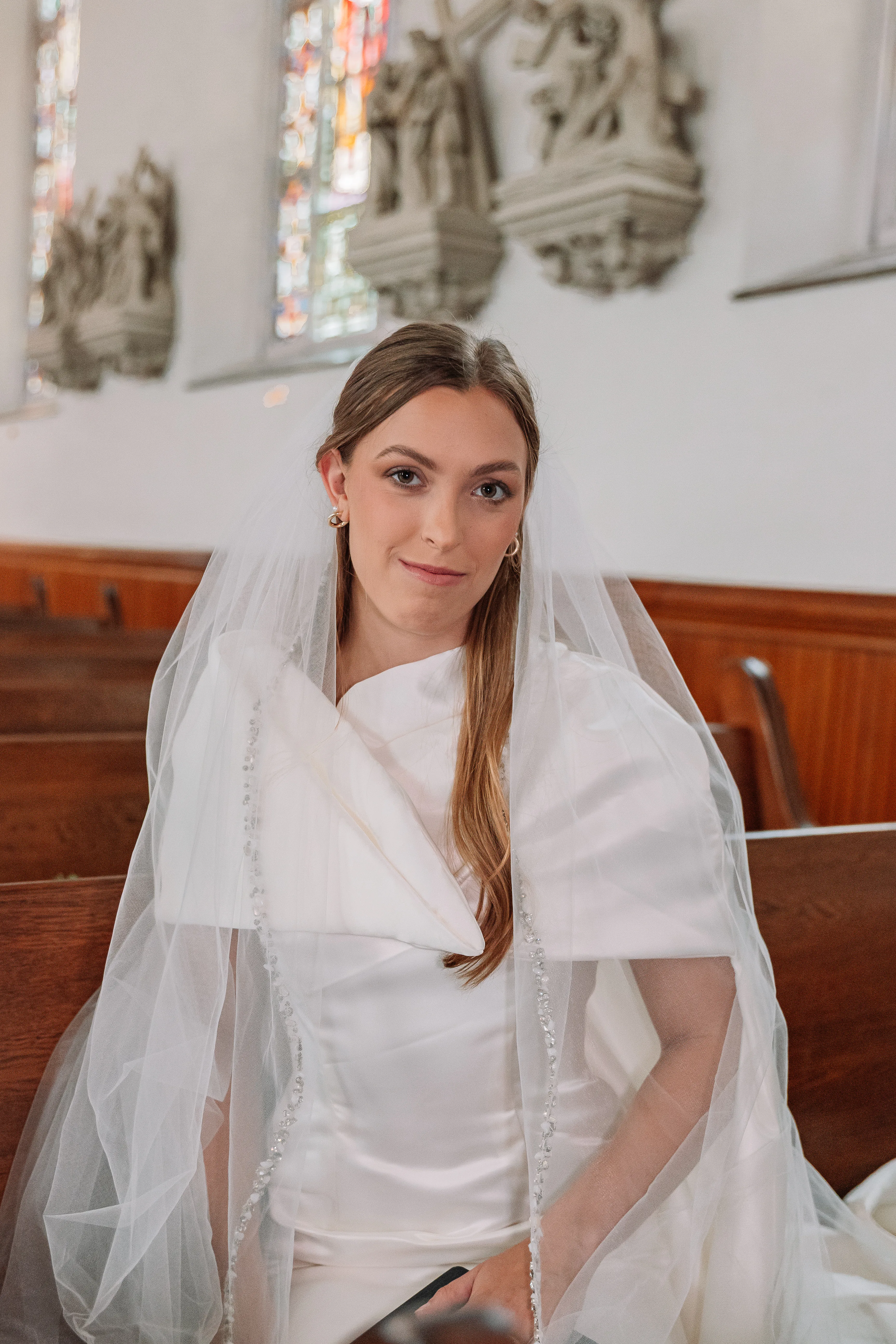 A seated bridal portrait showing the detachable cape dress beneath a veil in the church pews.
