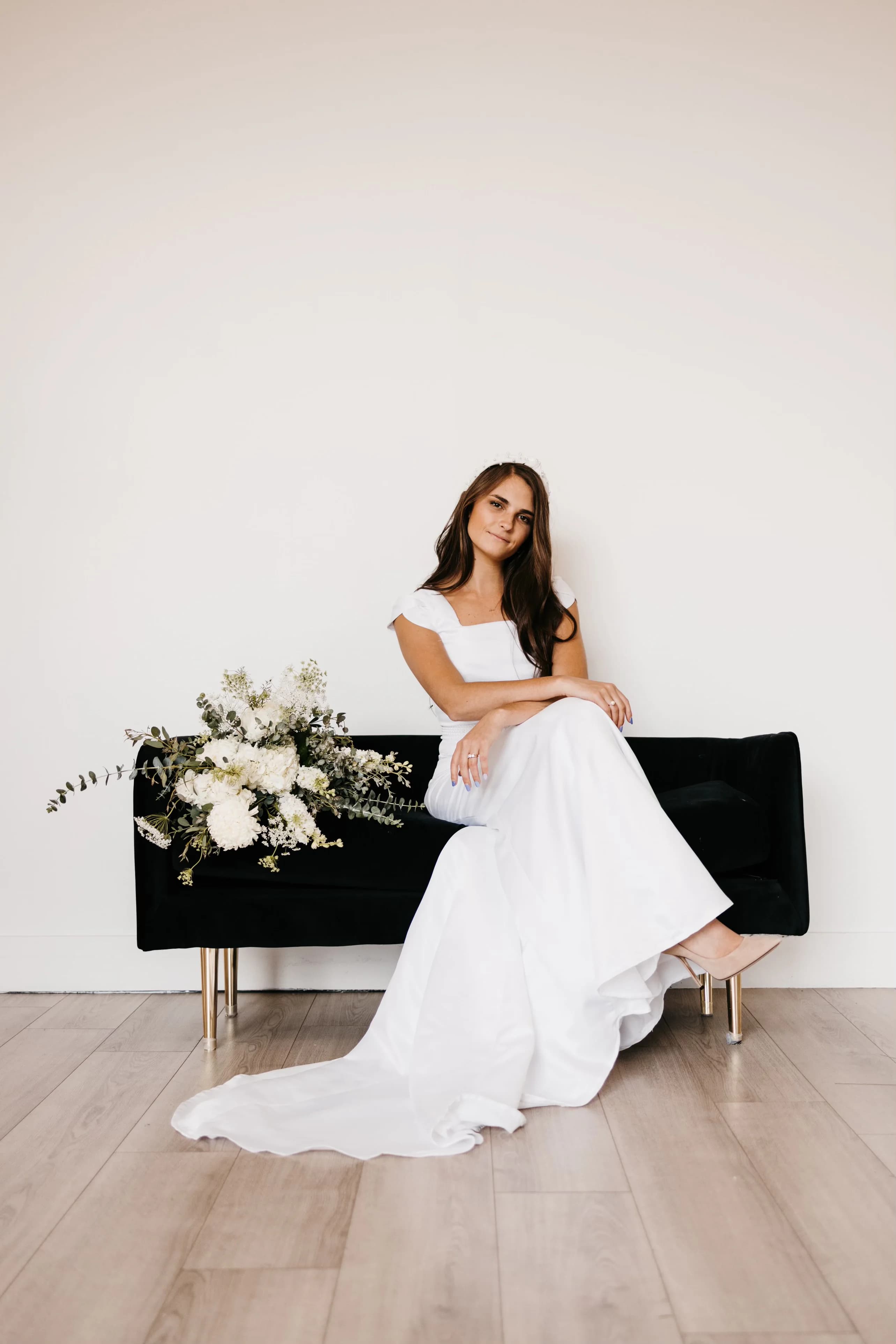 A seated bridal portrait on a black settee, showing the long skirt and soft square neckline of the first wedding dress Samantha Covey made.