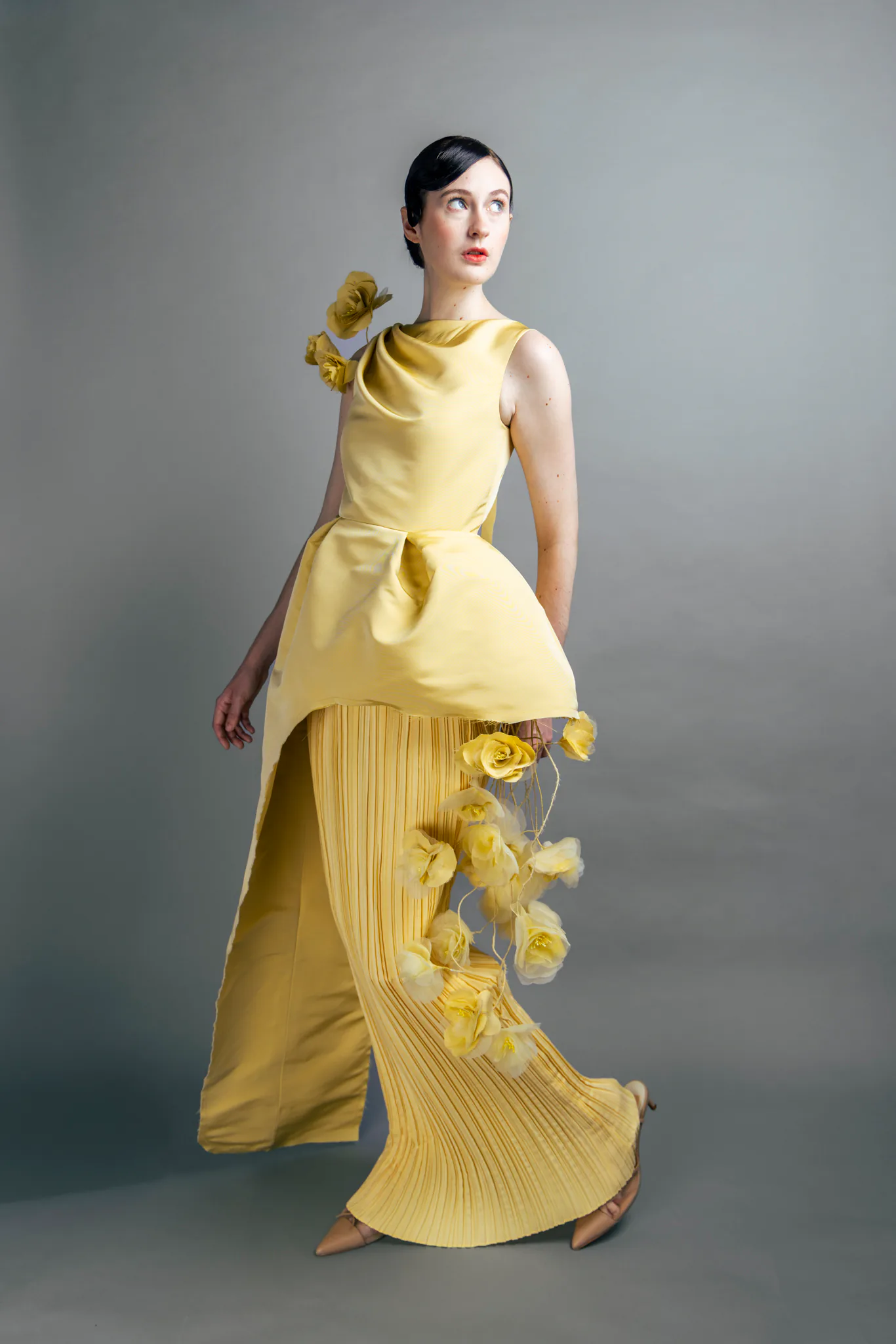 A full-length studio portrait of the yellow eveningwear look with pleated texture and floral embellishment.