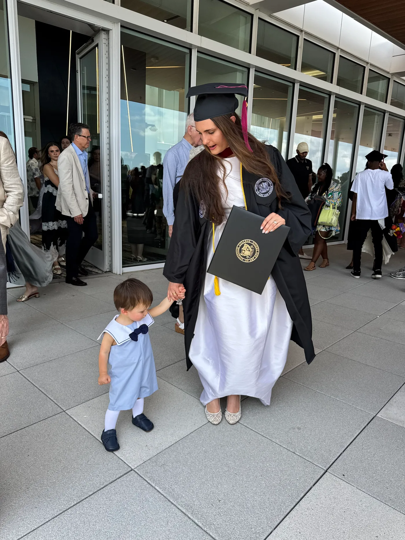 Samantha Covey in her SCAD graduation regalia holding her diploma beside a young child.