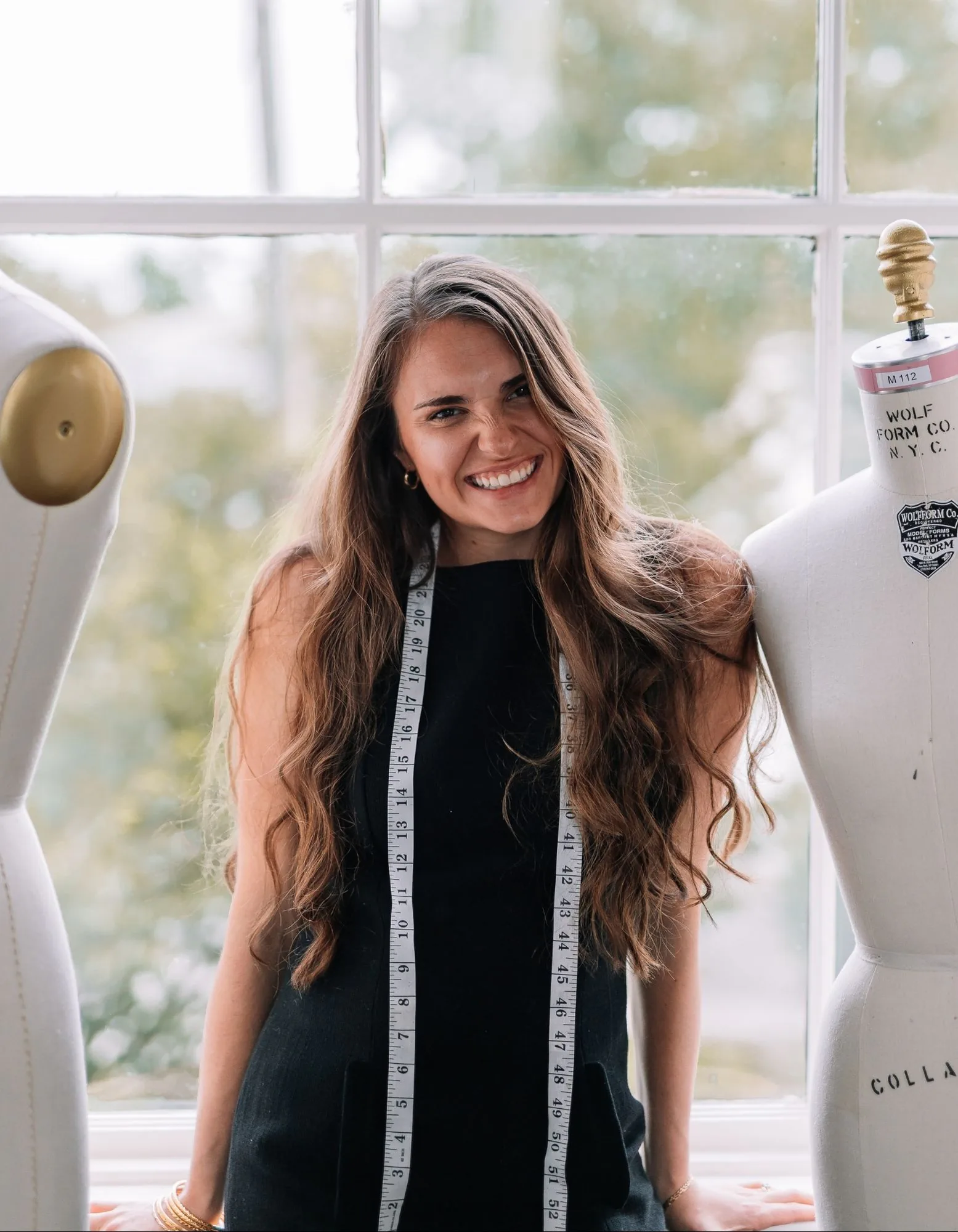 Samantha Covey smiling between dress forms in front of an atelier window.